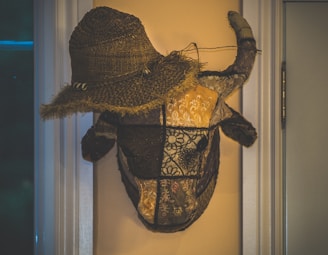 A decorative bull head mounted on the wall, adorned with a straw hat. The bull head appears to be made of patchwork fabric with intricate patterns, emphasizing a rustic, handmade aesthetic. The setting includes visible door frames and subtle lighting which casts soft shadows.