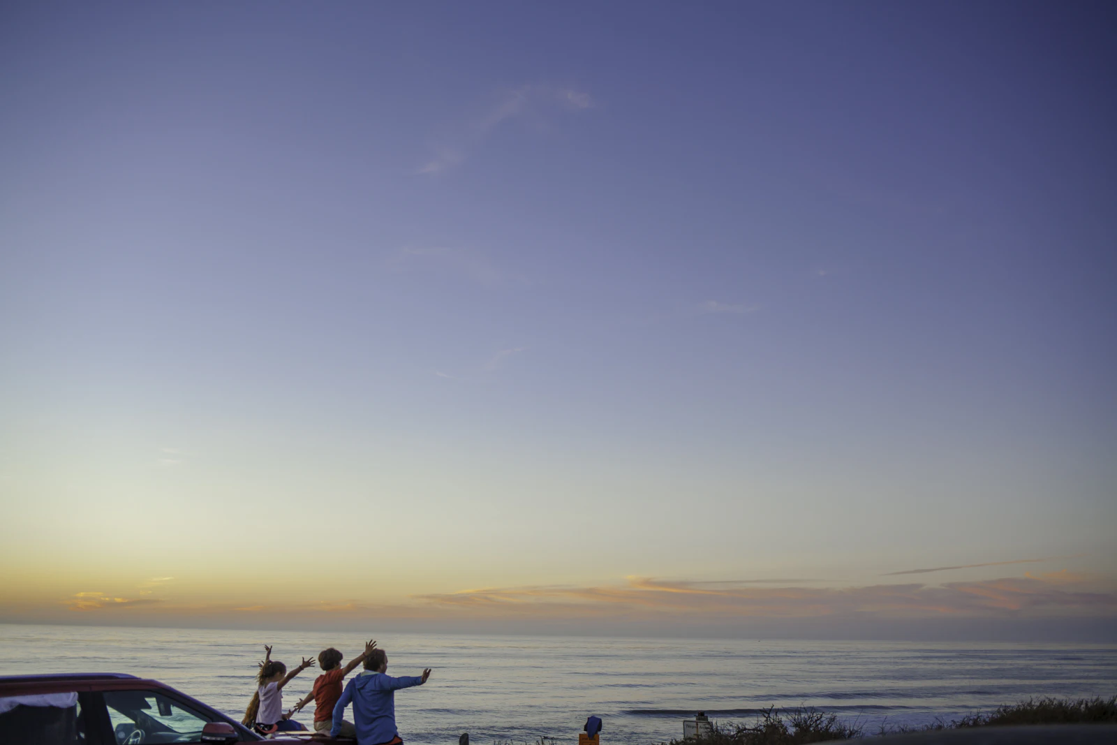 Family beside their car watching the sunset over the ocean on a road trip abroad