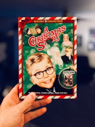 A hand is holding a DVD cover for a holiday movie titled 'A Christmas Story'. The cover features an illustration of a young boy wearing glasses, alongside several other characters in a festive theme with a red and green color scheme. The border is candy-cane striped, and the top reads 'Holiday Collection' with a red bow in the corner.