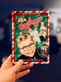 A hand is holding a DVD cover for a holiday movie titled 'A Christmas Story'. The cover features an illustration of a young boy wearing glasses, alongside several other characters in a festive theme with a red and green color scheme. The border is candy-cane striped, and the top reads 'Holiday Collection' with a red bow in the corner.