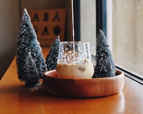 A glass of milk tea or coffee with ice cubes is placed in a wooden bowl on a wooden table. Liquid is being poured into the glass, creating a swirling effect. Surrounding the bowl are three small, artificial Christmas trees with white snow-like decorations. A window allows soft natural light to illuminate the scene, creating a cozy and warm atmosphere. In the background, a blurred sign with letters can be seen.