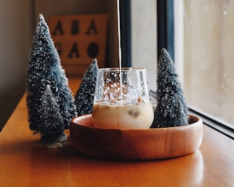 A glass of milk tea or coffee with ice cubes is placed in a wooden bowl on a wooden table. Liquid is being poured into the glass, creating a swirling effect. Surrounding the bowl are three small, artificial Christmas trees with white snow-like decorations. A window allows soft natural light to illuminate the scene, creating a cozy and warm atmosphere. In the background, a blurred sign with letters can be seen.