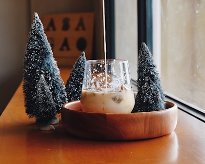 A glass of milk tea or coffee with ice cubes is placed in a wooden bowl on a wooden table. Liquid is being poured into the glass, creating a swirling effect. Surrounding the bowl are three small, artificial Christmas trees with white snow-like decorations. A window allows soft natural light to illuminate the scene, creating a cozy and warm atmosphere. In the background, a blurred sign with letters can be seen.