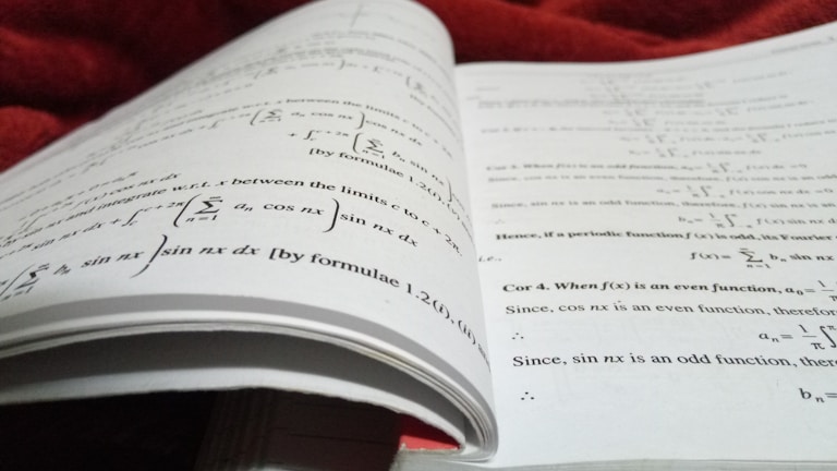 An open book with pages filled with mathematical formulas and text. The writing includes complex equations and mathematical symbols spread across two facing pages. The background is soft and out of focus, with what appears to be a red fabric.