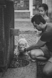 A man is kneeling beside a stone water fountain, helping a fluffy dog drink from the flow of water. A young girl stands nearby, watching the interaction with interest. The setting appears to be an outdoor area with cobblestone pavement and a blurred background.
