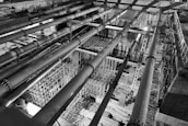 A black-and-white image of an industrial construction site featuring large horizontal pipes and intricate scaffolding structures. The perspective is from above, highlighting the complexity and scale of the construction work in progress. The lighting creates strong contrasts, adding depth and an architectural focus.