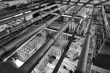A black-and-white image of an industrial construction site featuring large horizontal pipes and intricate scaffolding structures. The perspective is from above, highlighting the complexity and scale of the construction work in progress. The lighting creates strong contrasts, adding depth and an architectural focus.