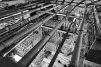 A black-and-white image of an industrial construction site featuring large horizontal pipes and intricate scaffolding structures. The perspective is from above, highlighting the complexity and scale of the construction work in progress. The lighting creates strong contrasts, adding depth and an architectural focus.