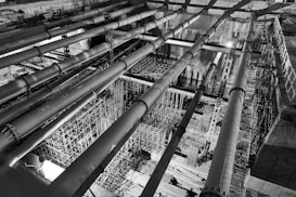 A black-and-white image of an industrial construction site featuring large horizontal pipes and intricate scaffolding structures. The perspective is from above, highlighting the complexity and scale of the construction work in progress. The lighting creates strong contrasts, adding depth and an architectural focus.