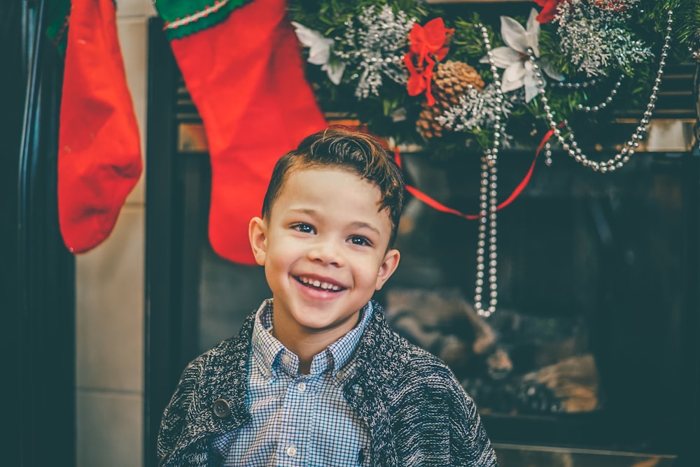 menino sorridente em camisa interna cinza e cardigan cinza em pé junto à lareira com decorações