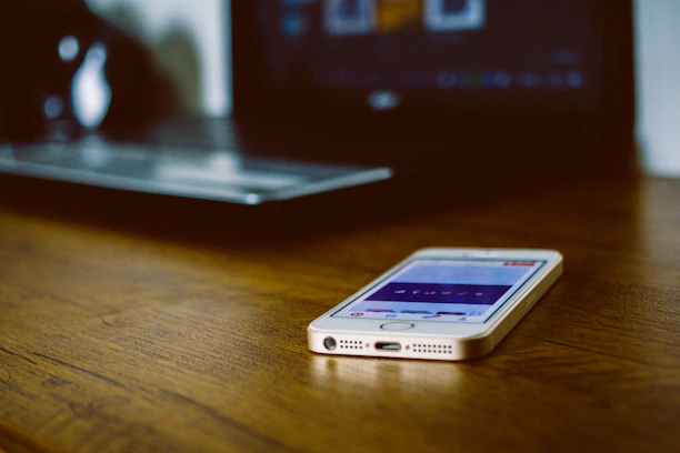 Close-up of a sleek smartphone resting on a wooden desk beside a laptop.