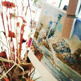 A close-up of a card with a watercolor-style illustration of a cityscape that includes modern skyscrapers and a body of water. The card is clipped to twigs or branches, surrounded by abstract, colorful shapes.