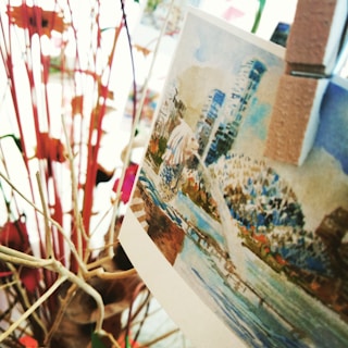 A close-up of a card with a watercolor-style illustration of a cityscape that includes modern skyscrapers and a body of water. The card is clipped to twigs or branches, surrounded by abstract, colorful shapes.