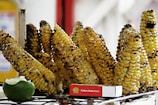 A collection of grilled corn cobs arranged in a row, their kernels charred and glistening. In front of the corn, there is a lime wedge and a small box labeled 'Palillos Doble Punta.' The background appears blurred with hints of red and white.