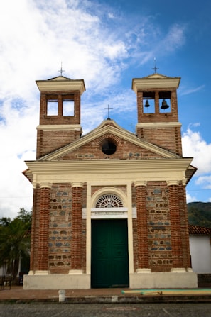 A historic brick church with two bell towers and a large green door. The building features a mix of brick and stone materials, adorned with a central white arch above the door. The sky is blue with scattered clouds, creating a serene backdrop.