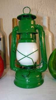 A green vintage lantern is situated on a white surface, surrounded by various colored bottles including gold, white, and red. The lantern has a glossy finish and features a white glass center.