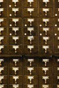 A set of vintage wooden library card catalog drawers with brass handles, labeled with different alphabetical ranges for organizing contents. The drawers are closely stacked, creating a pattern of repeated elements.
