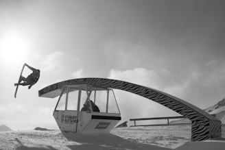 A snowboarder is performing an aerial trick near a ski lift structure with zebra print. The scene is set against a bright sky with the sun casting light over the snowy landscape. The structure has inscriptions including 'Stubai Glacier'.