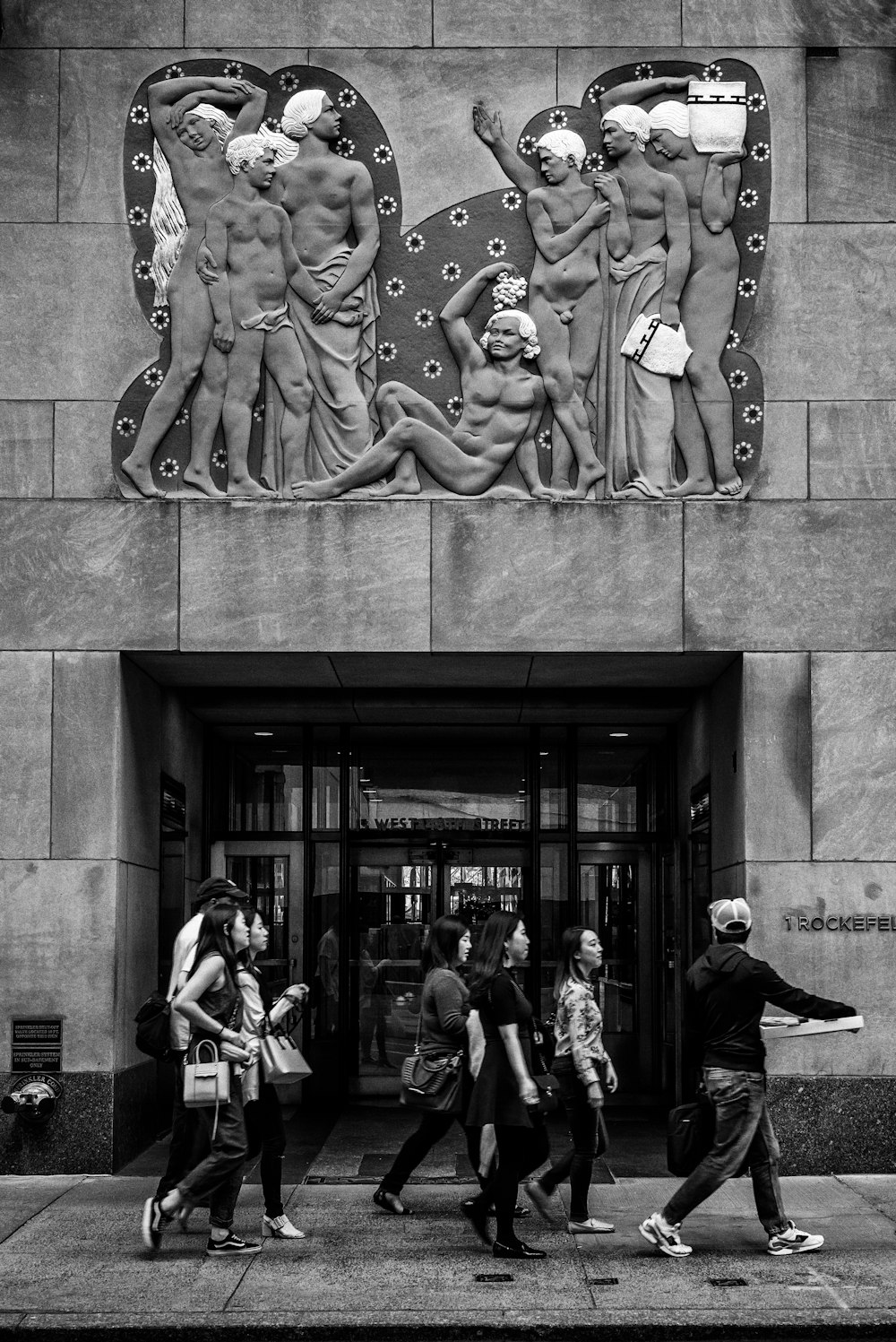 grayscale photography of people walking under sculpted wall