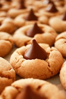 A close-up view of peanut butter cookies topped with a chocolate candy. The cookies have a soft, golden texture with a sugary coating, and each one is centered with a large, dark chocolate drop.