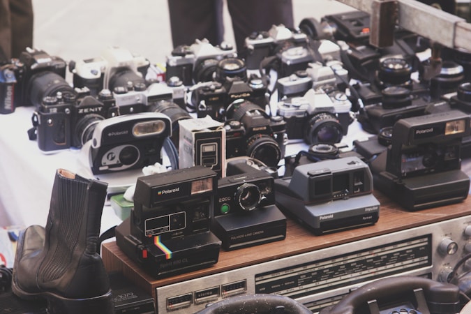 A collection of vintage cameras, including Polaroids, film cameras, and other analog equipment, is displayed on a table. The assortment features different models, some with lenses and others with their iconic designs visible. A pair of leather boots is partially visible in the foreground, and a stereo system can be seen at the bottom of the image.
