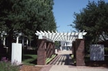 A wooden pergola structure supported by brick pillars is set in a landscaped area with trees and bushes. A walkway covered in shadows from the pergola leads towards residential buildings in the background. Flowers and trimmed bushes line both sides of the path.