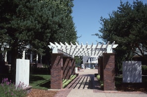 A wooden pergola structure supported by brick pillars is set in a landscaped area with trees and bushes. A walkway covered in shadows from the pergola leads towards residential buildings in the background. Flowers and trimmed bushes line both sides of the path.