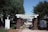 A wooden pergola structure supported by brick pillars is set in a landscaped area with trees and bushes. A walkway covered in shadows from the pergola leads towards residential buildings in the background. Flowers and trimmed bushes line both sides of the path.