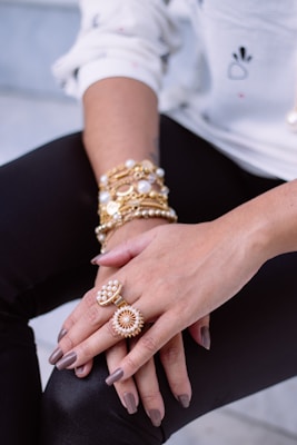 A close-up of a person's hands resting on their lap, adorned with multiple gold bracelets and rings, featuring pearl details. The person is wearing a white long-sleeve top with subtle patterns and black pants. The nails are painted in a muted taupe color.