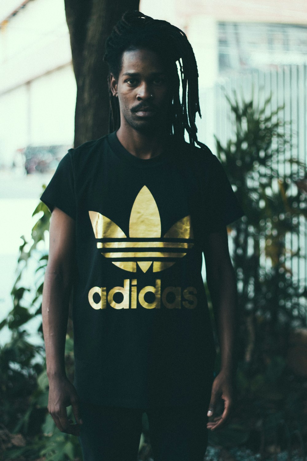 depth of field photography of man wearing black and gold adidas graphic crew-neck T-shirt standing in front of tree