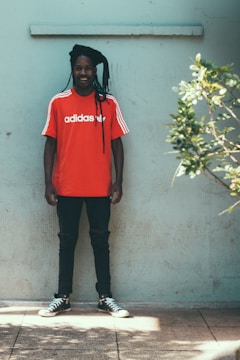 A person wearing a bright red Adidas t-shirt with white stripes on the shoulders stands against a light green wall. They are also wearing black jeans and black Converse-style shoes. The individual has long braided hair and is smiling. To the right, part of a leafy green plant is visible.