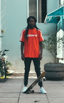 A person with long hair wearing a red Adidas shirt stands on a paved walkway holding a skateboard. The setting appears urban with a light-colored building and some greenery in the background.