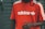 A person wearing a bright red Adidas t-shirt featuring white stripes on the shoulders and the brand's logo printed in white on the front. The background consists of a gray, metal roller shutter with some graffiti on the left side.