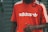 A person wearing a bright red Adidas t-shirt featuring white stripes on the shoulders and the brand's logo printed in white on the front. The background consists of a gray, metal roller shutter with some graffiti on the left side.