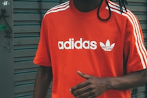 A person wearing a bright red Adidas t-shirt featuring white stripes on the shoulders and the brand's logo printed in white on the front. The background consists of a gray, metal roller shutter with some graffiti on the left side.
