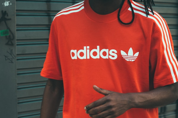 A person wearing a bright red Adidas t-shirt featuring white stripes on the shoulders and the brand's logo printed in white on the front. The background consists of a gray, metal roller shutter with some graffiti on the left side.