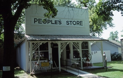 A quaint storefront features a white facade with a sign reading 'The People's Store.' The building has a rustic, country-style design with a porch and decorative trim. A small lawn surrounds it with trees providing shade. A vintage pump and a small horse ride are visible in the front.