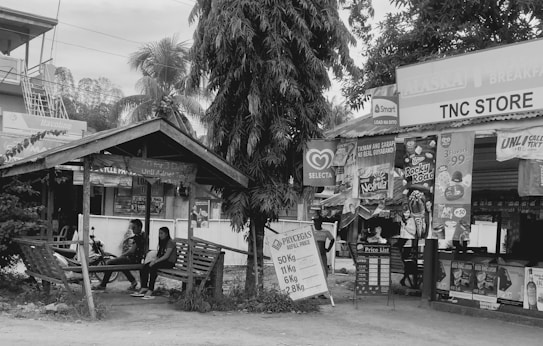 A small roadside store with a wooden shelter, featuring various advertising banners for different products like ice cream and mobile services. Two individuals are seated under the shelter, next to a parked motorcycle. The scene includes a large tree, another building in the background, and signboards displaying price lists.