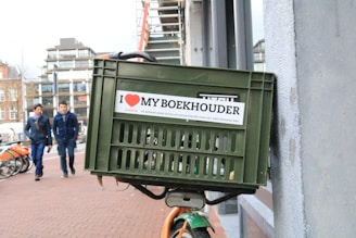 A green plastic crate is attached to the front of a bicycle, leaning against a wall on a city street. The crate features a sticker that reads 'I ❤️ MY BOEKHOUDER' with additional text in Dutch. In the background, two people walk on the sidewalk alongside parked bicycles, and several modern buildings are visible.