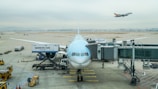 A large passenger airplane is stationed at an airport gate, preparing for boarding. The aircraft is connected to a jet bridge, with catering and loading vehicles positioned nearby. In the background, another airplane is taking off, soaring above the airport runway. The sky is overcast, creating a muted and calm atmosphere.