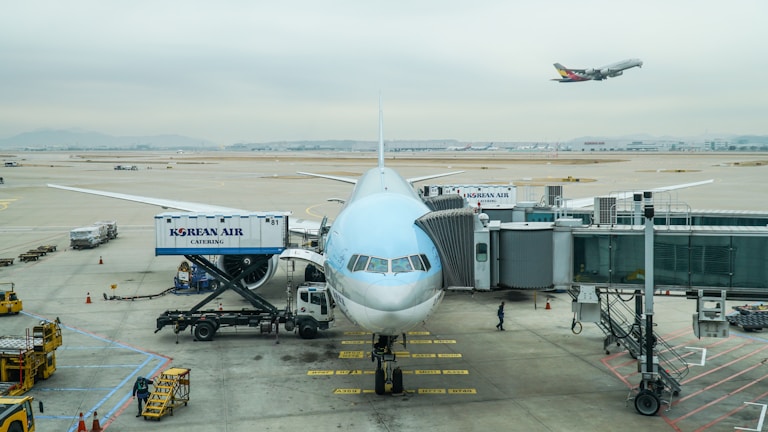 A large passenger airplane is stationed at an airport gate, preparing for boarding. The aircraft is connected to a jet bridge, with catering and loading vehicles positioned nearby. In the background, another airplane is taking off, soaring above the airport runway. The sky is overcast, creating a muted and calm atmosphere.