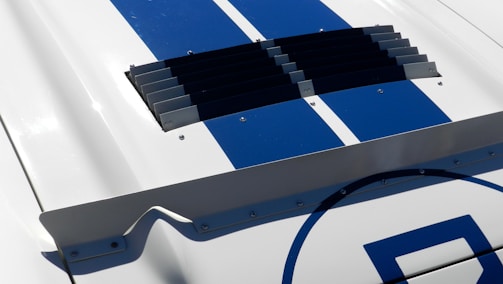 A close-up view of a section of a vehicle's body, featuring bold blue racing stripes on a white surface. There is a black vent with horizontal slats in the center, surrounded by rivets and a small aerodynamic spoiler attachment at the bottom of the image.