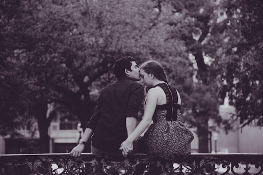 A couple sits on a wrought iron railing, holding hands. The man is gently kissing the woman on her forehead. They are surrounded by large, leafy trees and the setting appears still and serene. The image is in black and white, adding a timeless and romantic feel to the scene.