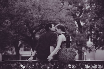 A couple sits on a wrought iron railing, holding hands. The man is gently kissing the woman on her forehead. They are surrounded by large, leafy trees and the setting appears still and serene. The image is in black and white, adding a timeless and romantic feel to the scene.