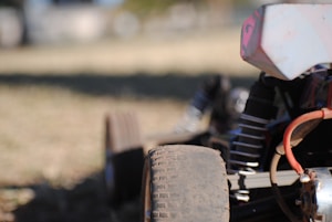 The foreground features part of a rugged remote-controlled car with prominent dirt-covered wheels and visible engine components. A coil spring and a portion of the chassis are apparent. The background is blurred, suggesting an outdoor setting with grassy terrain.