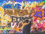 A vibrant wall covered in colorful graffiti, with phrases like 'Love Yourself First' and 'Yes' prominently featured. A person stands in front of the wall, appearing to add to the artwork with a spray can.