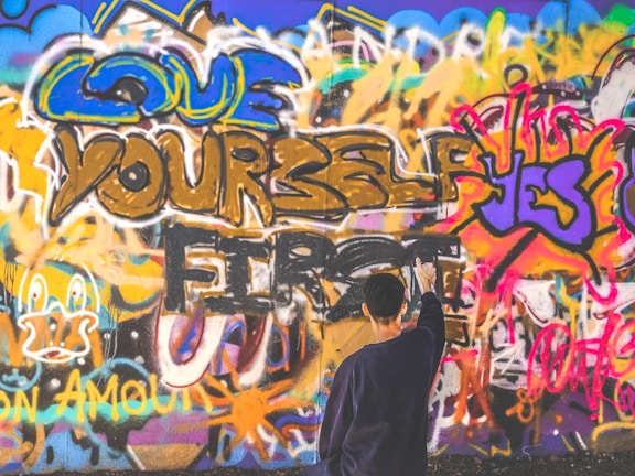 A vibrant wall covered in colorful graffiti, with phrases like 'Love Yourself First' and 'Yes' prominently featured. A person stands in front of the wall, appearing to add to the artwork with a spray can.