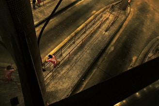 A person wearing a red sports jersey is standing on a pedestrian walkway, possibly in an urban area. The scene is captured from above at an angle, highlighting the geometric patterns of the tiled pavement and the metal railings. The lighting is dim and has a yellowish hue, casting shadows across the walkway.