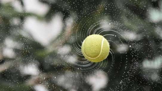 A tennis ball is caught mid-air with droplets of water spiraling around it, creating a dynamic and energetic effect. The background is blurred with natural greenery providing a contrast to the vivid yellow of the ball.