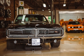 A vintage black muscle car is prominently displayed in a showroom setting. Its glossy exterior and classic grille are highlighted under indoor lighting. To the left, there are several new rims mounted on the wall. In the background stands an orange modern sports car, contrasting with the classic design of the black car.